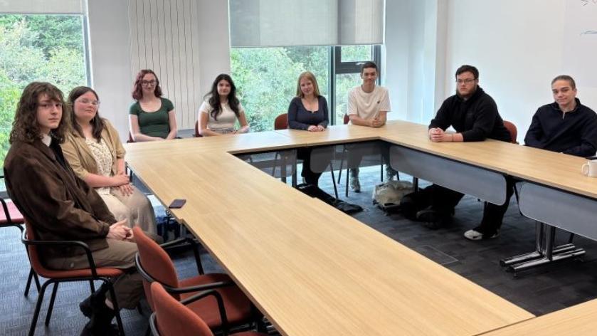 Eight classmates sat around a rectangular formation of chairs and tables