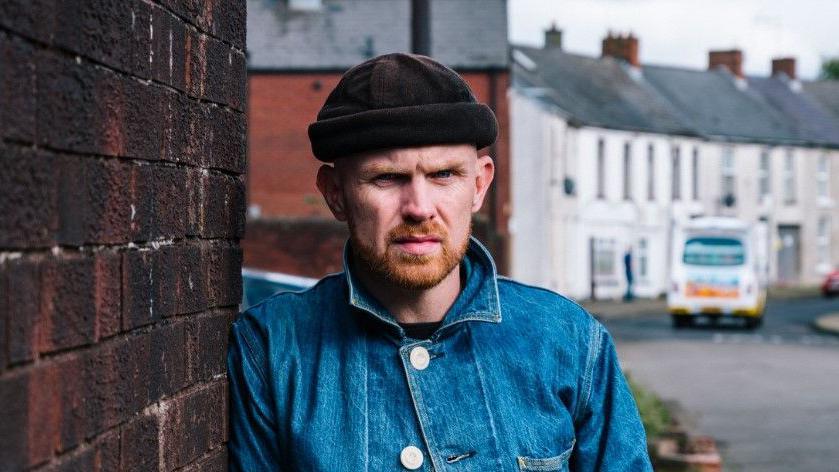 Rory Carson wearing a blue denim jacket with the collar up. He is also wearing a grey skull cap. He is looking directly at the camera with a serious expression while leaning against a red brick wall in a street. There is an ice-cream van in the background