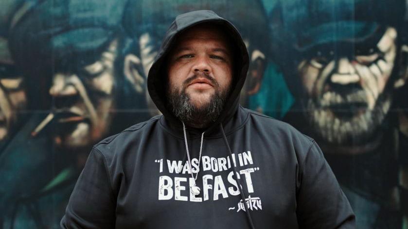 Jun Tzu. He has a dark beard, wearing a black hoodie with the hood up, on the front of it it reads 'I Was Born In Belfast'. A mural of men wearing flat caps and smoking cigarettes is behind him.
