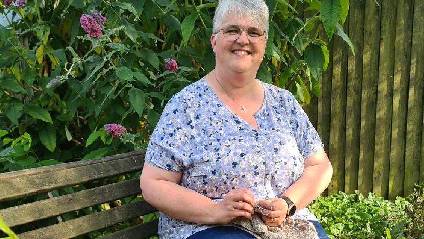 Sue Bailey, with glasses and grey short hair and wearing a blue top, sitting on a bench, smiling and crocheting