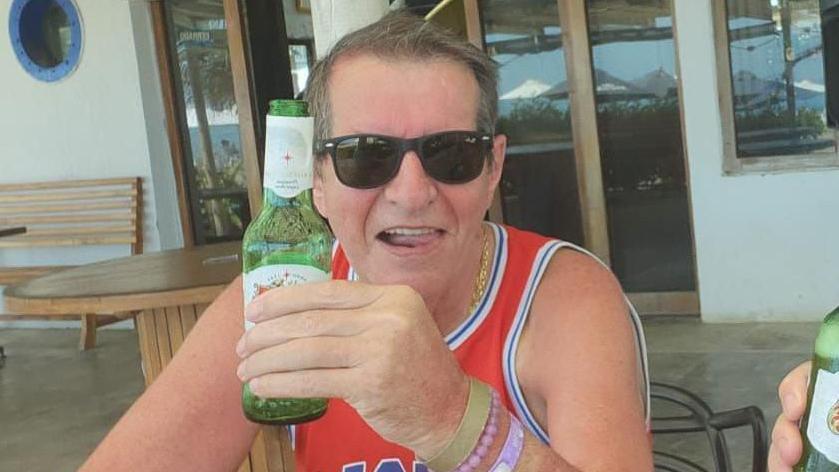 Paul Longworth sat at a table with a bottle of beer. He is smiling and has on dark sunglasses. 
