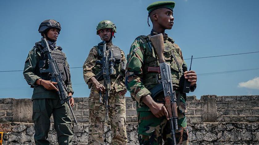 Three men in military fatigues and holding rifles stand in front of a breeze block wall.