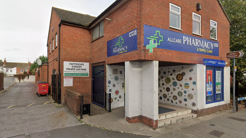 A red brick building with two blue banners reading Allcare Pharmacy and Travel Clinic on either side of the building in view in the picture