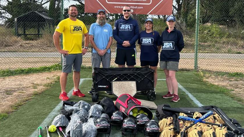 Three men and two women from the Jersey Softball Association stand behind a load of softball equipment. There are stood in a batting cage with Jersey Softball Association banners of the fencing.