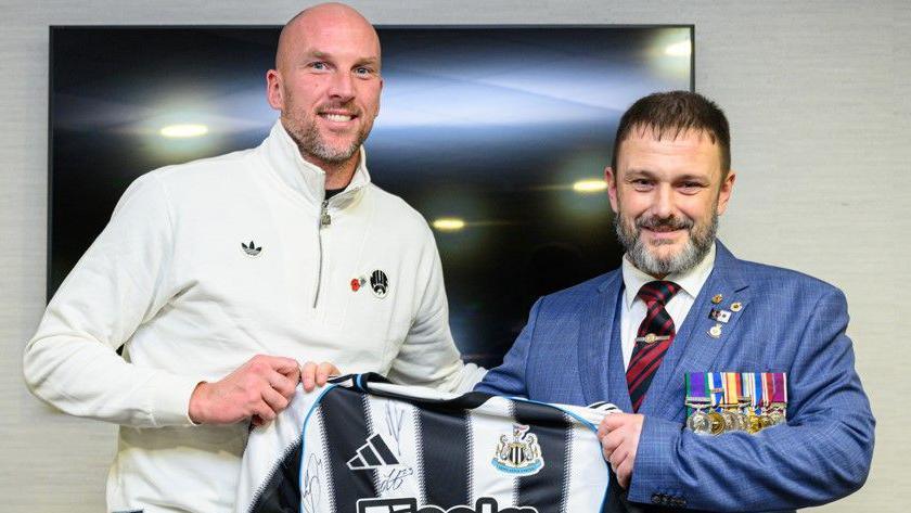 John Ruddy standing next to an armed forces veteran. The man is wearing a blue suit and a seven medals. They are both holding between them a signed Newcastle United shirt.