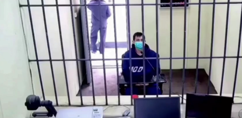 A man sitting in a hoodie and a surgical mask behind a metal grille in a room. There are two computer screens on a desk in front of the grille. Behind him the door is open and a person is standing outside the door.