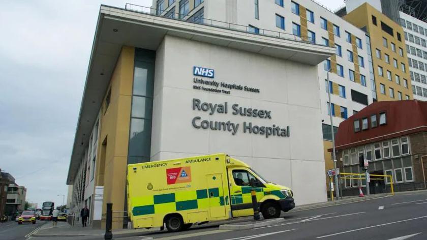 A ambulance driving up a hill past a large cream building with the words Royal Sussex County Hospital on it