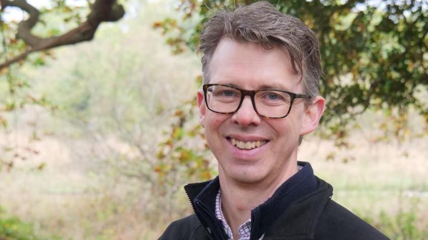 Eliot Lyne has light brown hair, and wears tortoise shell glasses. He is smiling, standing in a woodland setting. He is wearing a shirt with a squared lines pattern and has a black fleece over that.