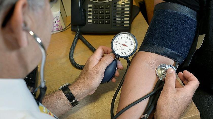 A close up of a doctor taking a patient's blood pressure. The patient has his arm on a desk, and the doctor is pressing the device against his arm. 