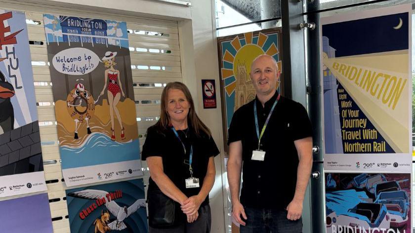 Two adults stand beside a wall of colourful railway posters