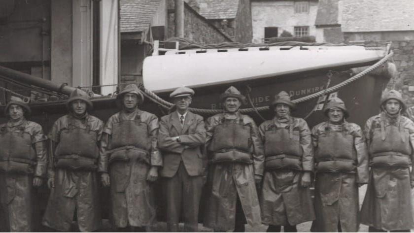 A black and white image of men standing in front of the vessel. The men are lined up in a row and are covering the vessel's name. They are wearing hats, life jackets and long coats.