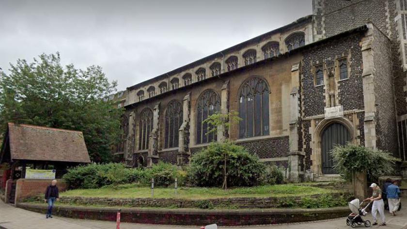 A street view of St Andrew's Church which has dark flints and five large stained windows, with another row of smaller windows above. There are some shrubs and grass in front of the church and a small wall bordering the corner path.