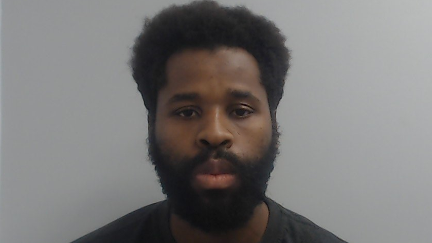 Police custody mugshot of Fabrice Kapena, who has a black beard and is wearing a black t-shirt.