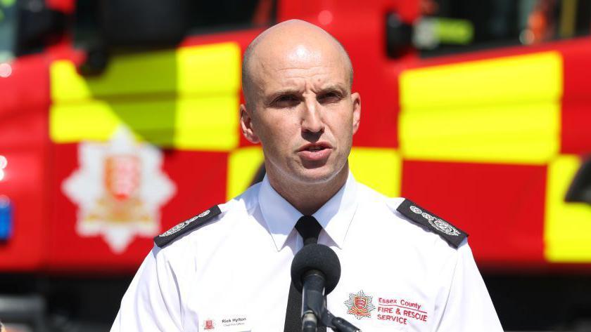Rick Hylton delivers a statement from behind a podium. He is in a white fire service shirt and is standing in front of a fire engine. 