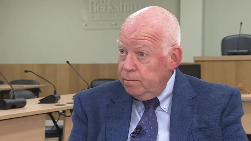 Picture of West Berkshire Council leader Jeff Brooks sitting in the Council chamber. With a serious expression he's wearing a blue suit, blue shirt and a blue tie
