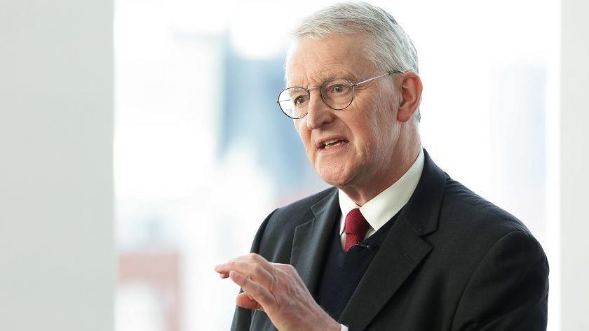 Hilary Benn has white hair and light framed glasses and is talking with his hand slightly raised. He is wearing a dark suit, dark v-neck jumper, white shirt and red tie.