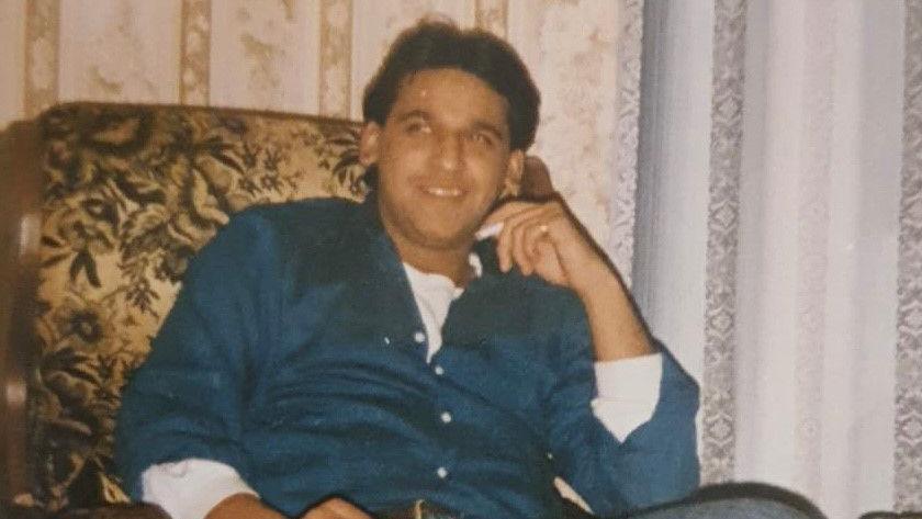 Shami wearing a blue shirt with white buttons and a white top underneath. He is sitting on a patterned arm chair