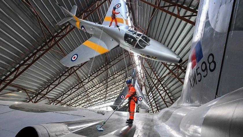 Two people, dressed in all orange and wearing helmets, attached to ropes are cleaning two RAF planes hanging from the ceiling of a hangar.