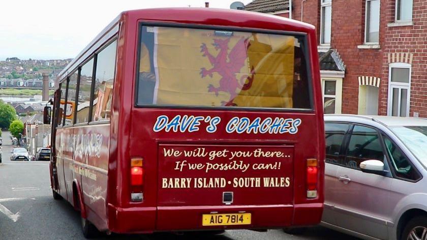 A red coach with blue writing saying Dave's Coaches. It has the Somerset flag displayed across the rear window.