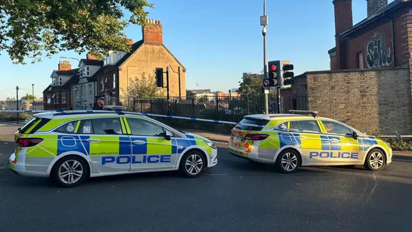 Two police cars in front of a police cordon blocking a road in Ipswich. A row of terraced houses can be seen in the background. 