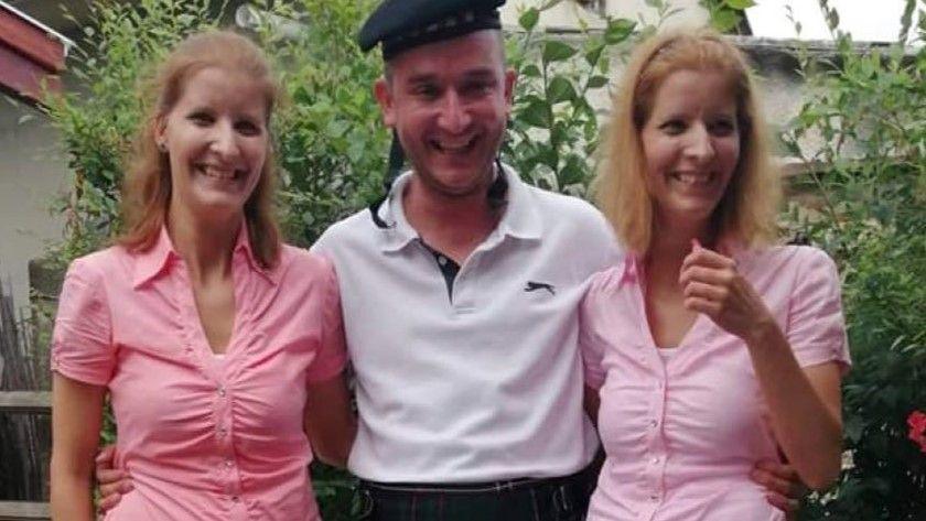 Twin sisters Eliza and Henrietta Huszti in pink tops photographed either side of their brother, Jozsef, who is wearing a white top. They are all smiling.