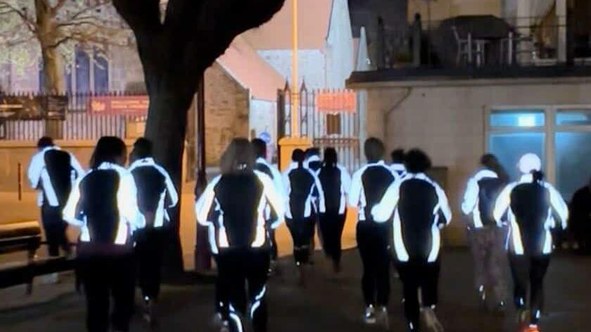A group of women running in a group in the dark. They are wearing hi-vis jackets and running on a street.