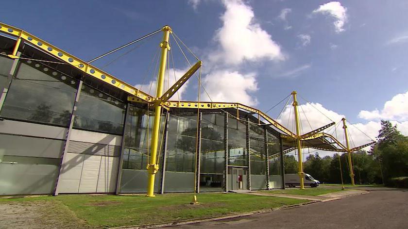A shot of part of an unusual warehouse under a blue sky. Three areas of pitched roof can be seen, with big glass panels, but the structural metal beams are painted bright yellow and are on the outside, not hidden.