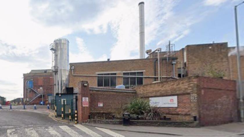 A brick building with metal structures including a tall cylindrical tank and a chimney. In the foreground is a zebra crossing leading up to a gated entrance.