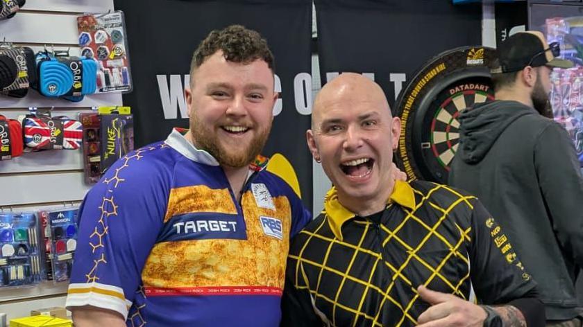 Two men standing side-by-side. One has Short dark curly hair and ginger facial hair. He is wearing a blue sports top with patterns. Next to him is a man with a bald head and a black top with a yellow pattern. Behind them are darts accessories.