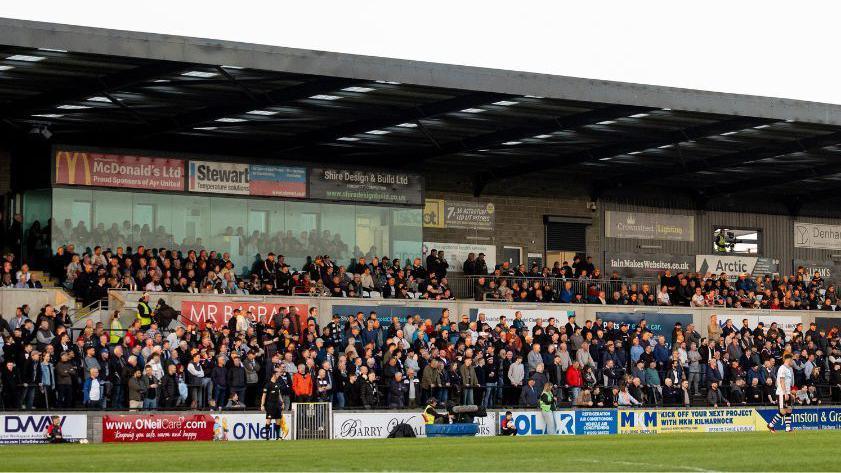 A general view of a stand at Somerset Park. Hundreds of fans are tightly packed into a stand covered in advertising hoardings. There are some players and match officials in the foreground on the pitch.