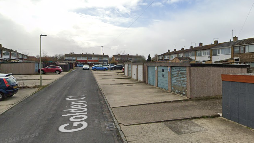 Golden Close in Tuffley, which is a residential area, during the day time. There are 7 garages to the right with a row of terraced houses behind them. 