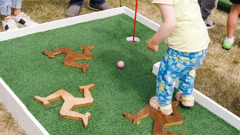 A toddler plays crazy golf. They're standing on a putting green, with artificial grass, which has wooden triskelion's. They are wearing flowery crocs, and blue flowery jeans with a yellow t-shirt.