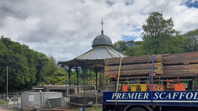 Scaffolding being erected at the fenced-off bandstand. A lorry with building materials is parked in front of it. The structure has ornamented railings and a grey roof with a cupola on top of it. It is surrounded by trees.