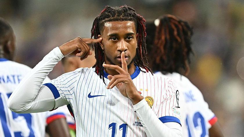 Bayern Munich and France forward Michael Olise with his right hand pointing at his temple and his left index finger over his lips