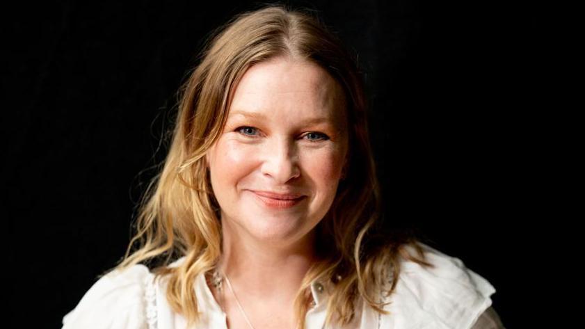 Joanna Page, a blonde woman looking at the camera and smiling. Her shoulder-length blonde hair is untied and she has a white top on. It is a head and shoulders shot of her. And she is in front of a black background.
