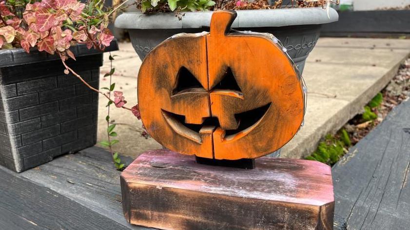 A small pumpkin carved out of wood and sprayed with orange paint. It's sat in front of two planters on a garden patio.