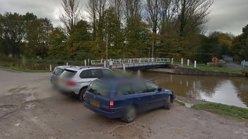 There are three cars parked at the side of a canal which has a bridge over it