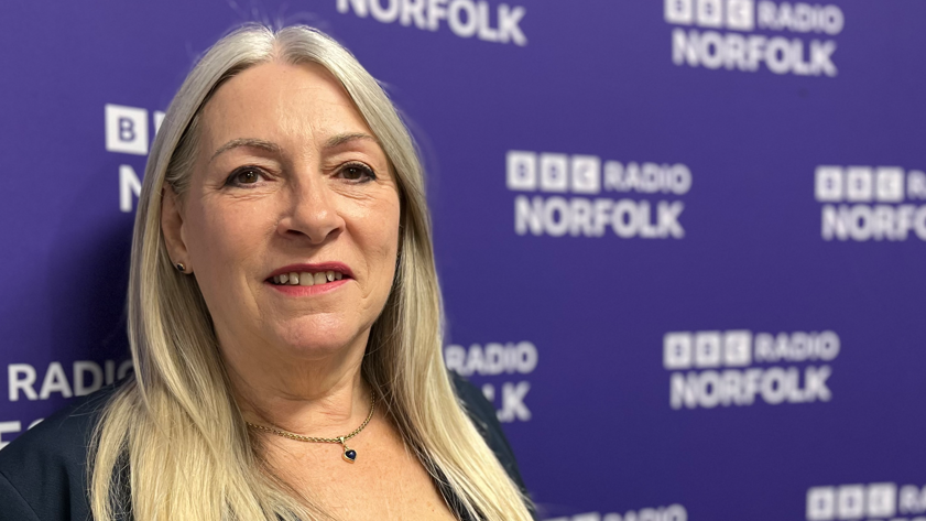 Kay Mason Billig standing in front of dark blue wall with BBC Radio Norfolk across it repeatedly in white capital letters 