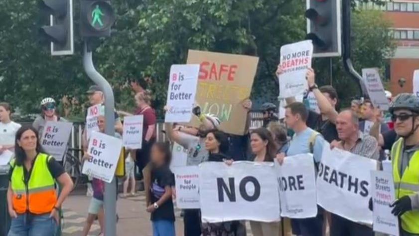 People gathered at a protest next to some traffic lights at a pedestrian crossing. People are holding signs calling for safer streets in Birmingham.