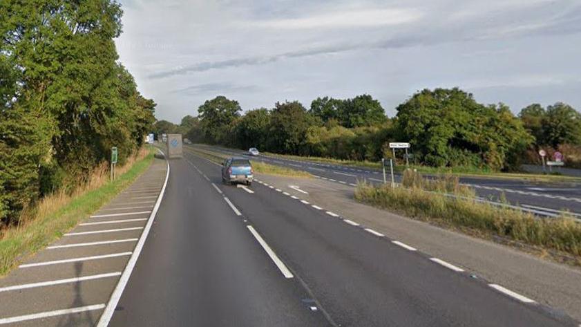 The A1 road going southbound with the northbound carriage on the right side. It is lined by trees on both sides and there is a junction off to the right. There is a sign saying "Water Newton" on the right.