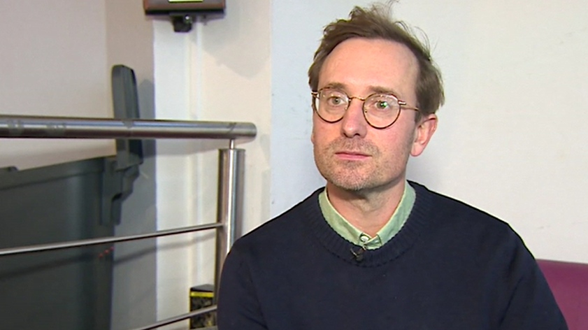 A man with short brown hair and round glasses. He is wearing a dark blue jumper and is sat on a sofa next to some metal railings.