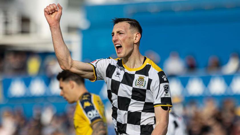 Robert Bozenik in a black-and-white checked Boavista shirt celebrates a goal by putting his right fist up above his head