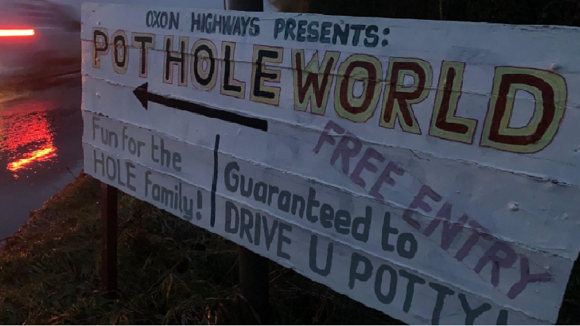 A sign reading: "Oxon Highways presents: Pothole World", "fun for the hole family" and "guaranteed to drive you potty". It is right next to a road.