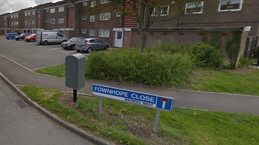 A road sign at the junction of Fownhope Close. There is a grass verge, with a row of houses in the background, and parked vehicles on driveways in front of them.