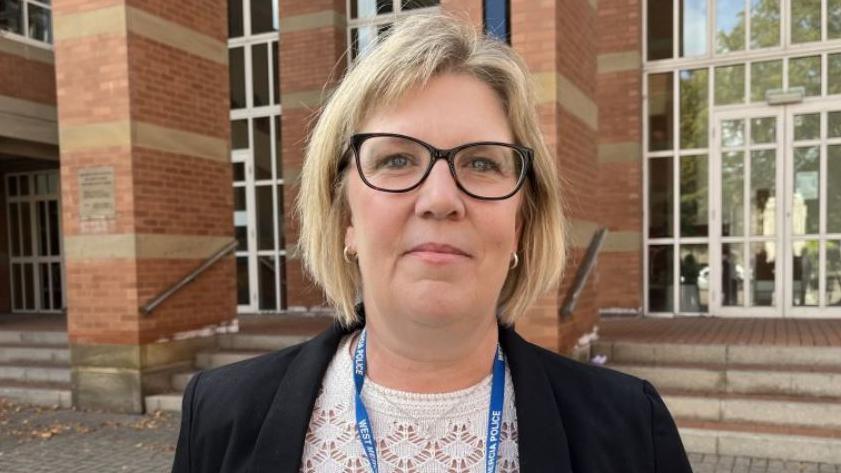 A woman with blonde hair, a dark jacket, white top and a blue lanyard on the steps of a brown brick building