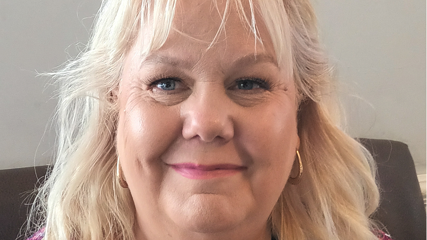 A woman in her 50s with long blonde hair and blue eyes. She is wearing gold hooped hearings and is smiling.