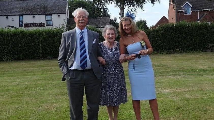 David Bays pictured with his wife Janet and daughter Rachel at a wedding in 2017. The three are standing outside on a lawn, with the Five Bells pub in the background behind a hedge. David has white hair and a moustache and is wearing glasses. He is wearing a grey suit, white shirt and blue-striped tie. Janet is wearing a blue dress with white dots and has a long pearl necklace. Rachel is wearing a light blue strapless dress and blue fascinator, and is holding a camera.