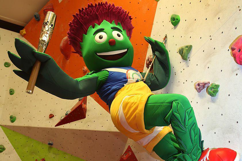 Clyde the mascot, an animated thistle, with green skin styled like leaves and purple spiking hair wearing a Glasgow 2014 sport outfit. It is hanging off a climbing wall holding the 2014 commonwealth games baton.