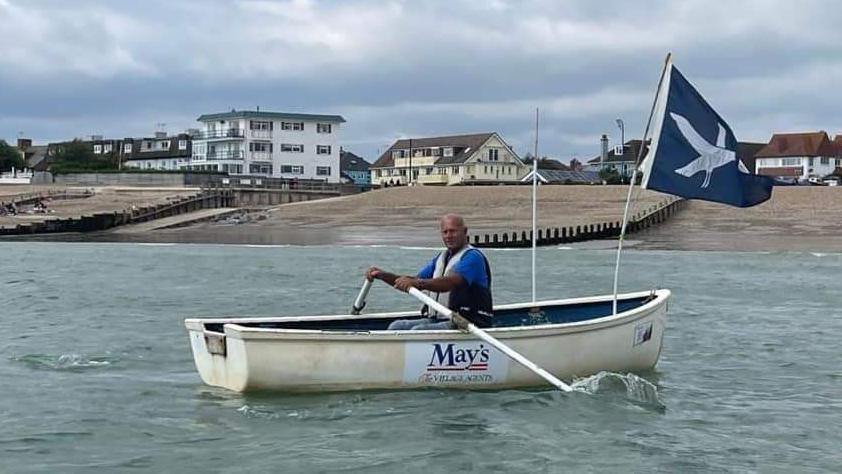 A man wearing a blue shirt and white life jacket rowing a white dinghy boat. The boat is flying a blue flag with a white dove on it. The boat is in the sea with the beach and houses in the background