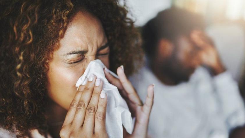 A Woman with curly hair is sneezing into a tissue. There's a man in the blurred background behind the woman who has his head in his hands. 